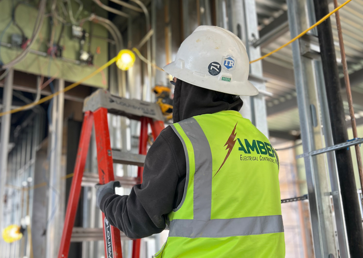 Electrician in safety gear working on electrical installation, Amber Electrical branding visible on vest, construction site background with tools and wiring.